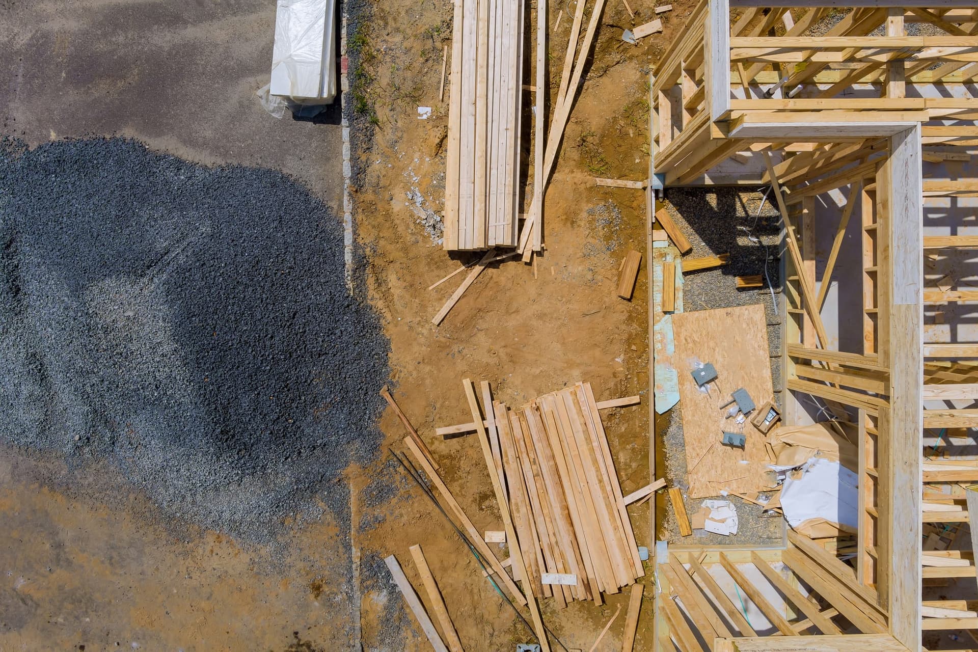 Aerial view of a residential construction site with stacked lumber and a pile of aggregate beside a partly framed house