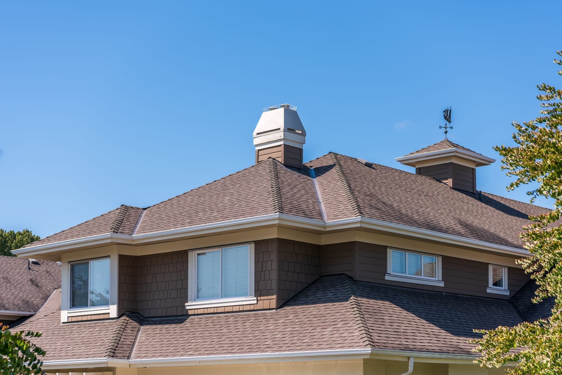 A hipped asphalt-shingle roof with a brick chimney, cupola and weather vane against a clear blue sky