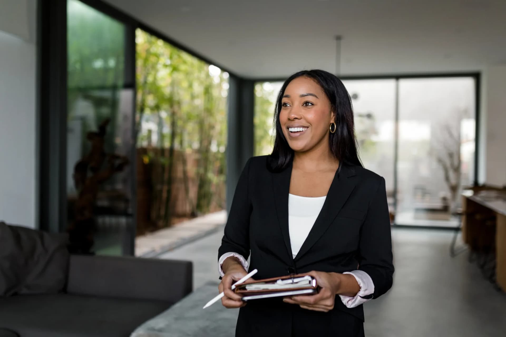 A real estate agent holding a tablet, standing in a bright modern living space with floor-to-ceiling glass